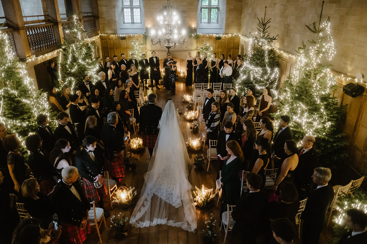 people assembled for a wedding ceremony in the ballroom at Achnagairn Castle. It is decorated with fairylights and Christmas trees. A bride walks down the aisle on the arms of a man in a kilt.