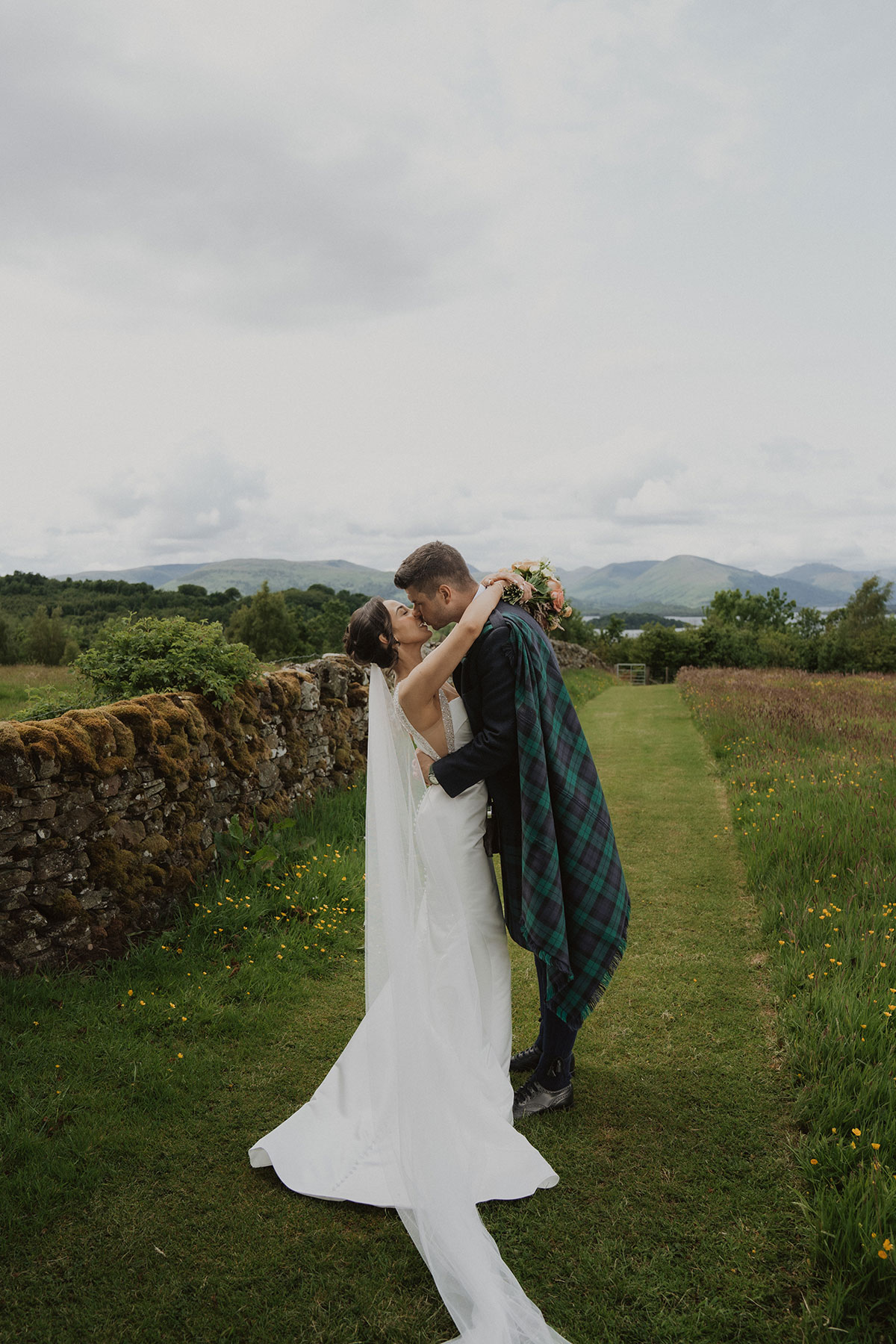 Bride and groom kiss beside moss-covered stone wall with wildflowers and countryside views in Scotland