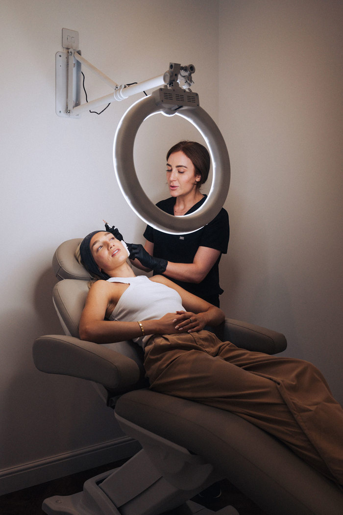 a woman lies down in a treatment chair as a large ring light shines on her face and a technician uses gloves to apply something to the client's face