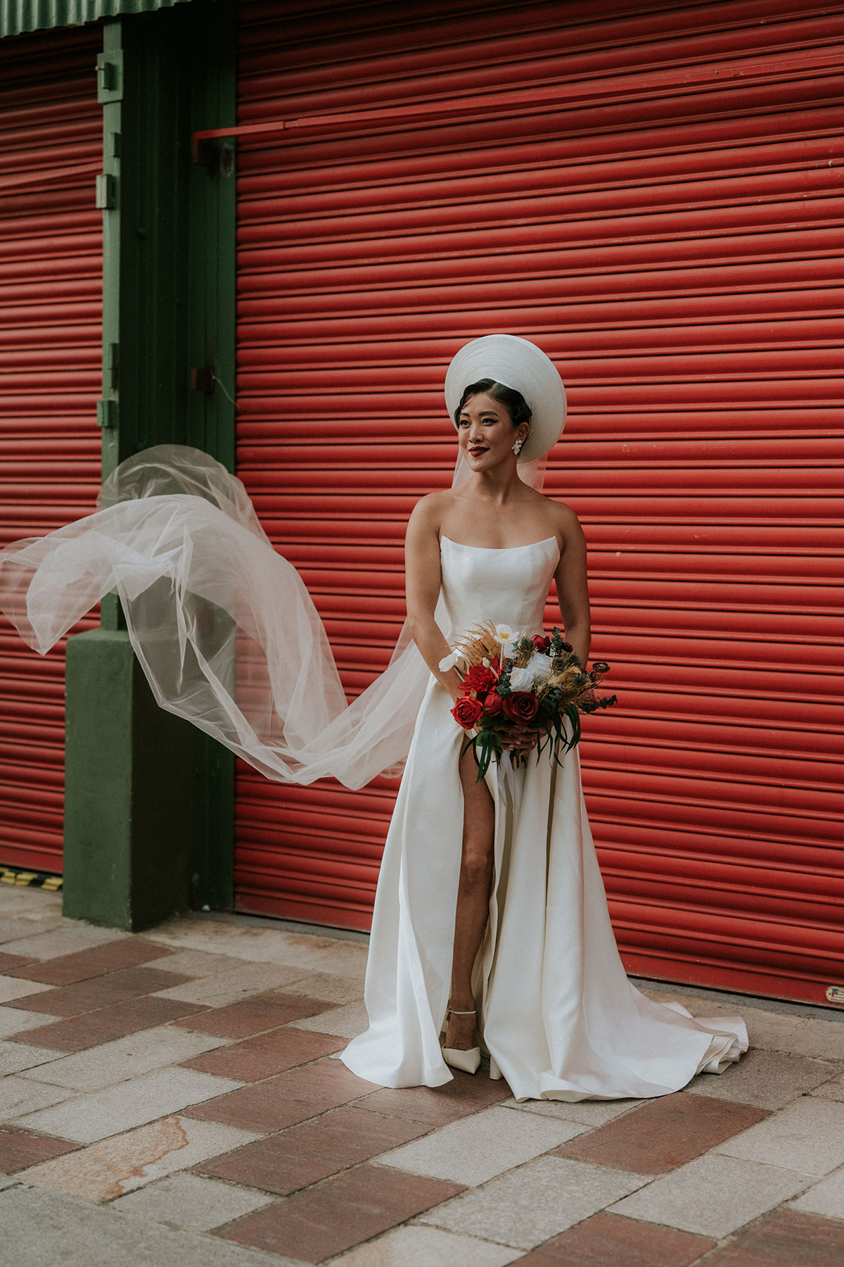 Bride in strapless white wedding dress with Vietnamese headpiece holding bouquet against red shutter at BAaD Glasgow