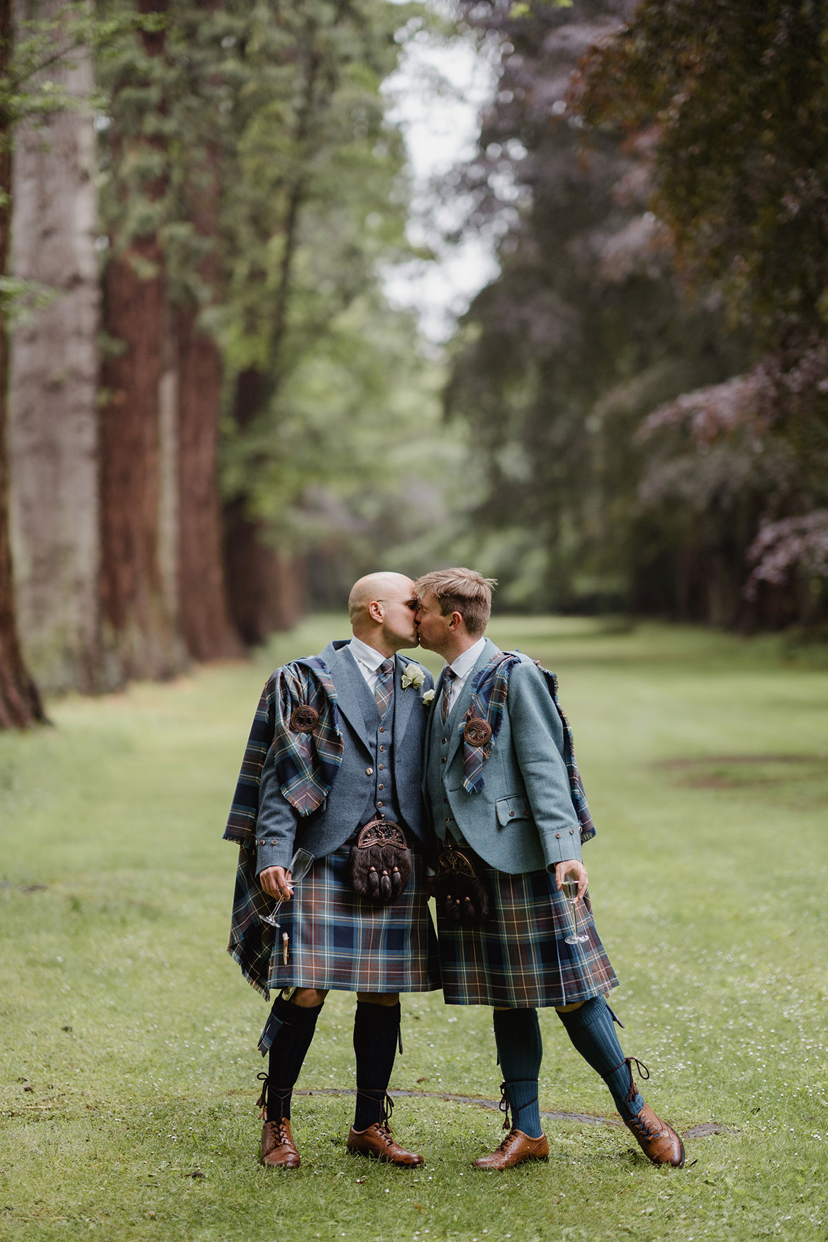 The couple kiss beneath tall trees in a woodland setting, both wearing coordinating blue tartan kilts and jackets