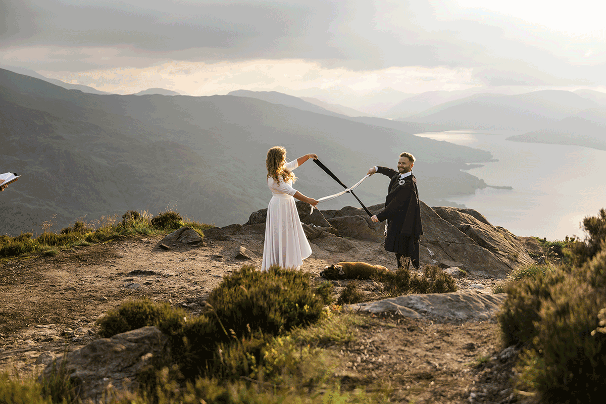 Bride and groom take part in a handfasting ritual during an outdoor Scottish elopement, surrounded by dramatic mountain and loch scenery