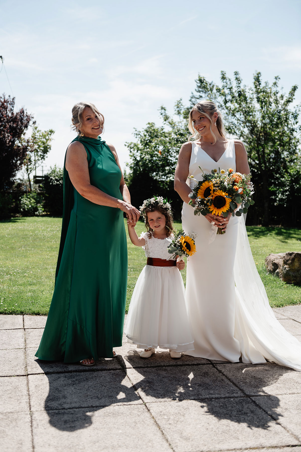 Bride, bridesmaid in green dress and young flower girl holding sunflower bouquets standing outside on a sunny day