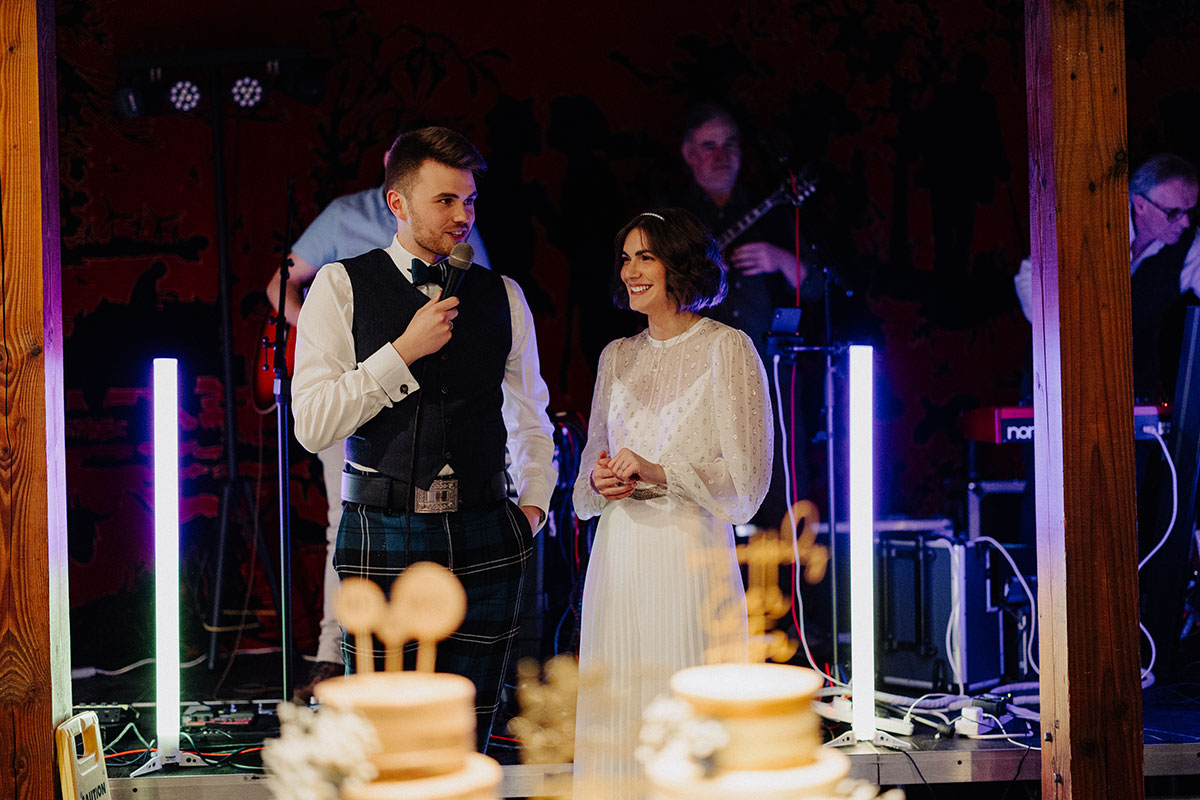 Bride and groom standing together as groom gives a speech