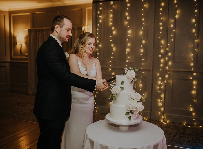 A bride and groom cutting into their three tier cake which has white roses on it and is on a small table in front of a wall of fairylights