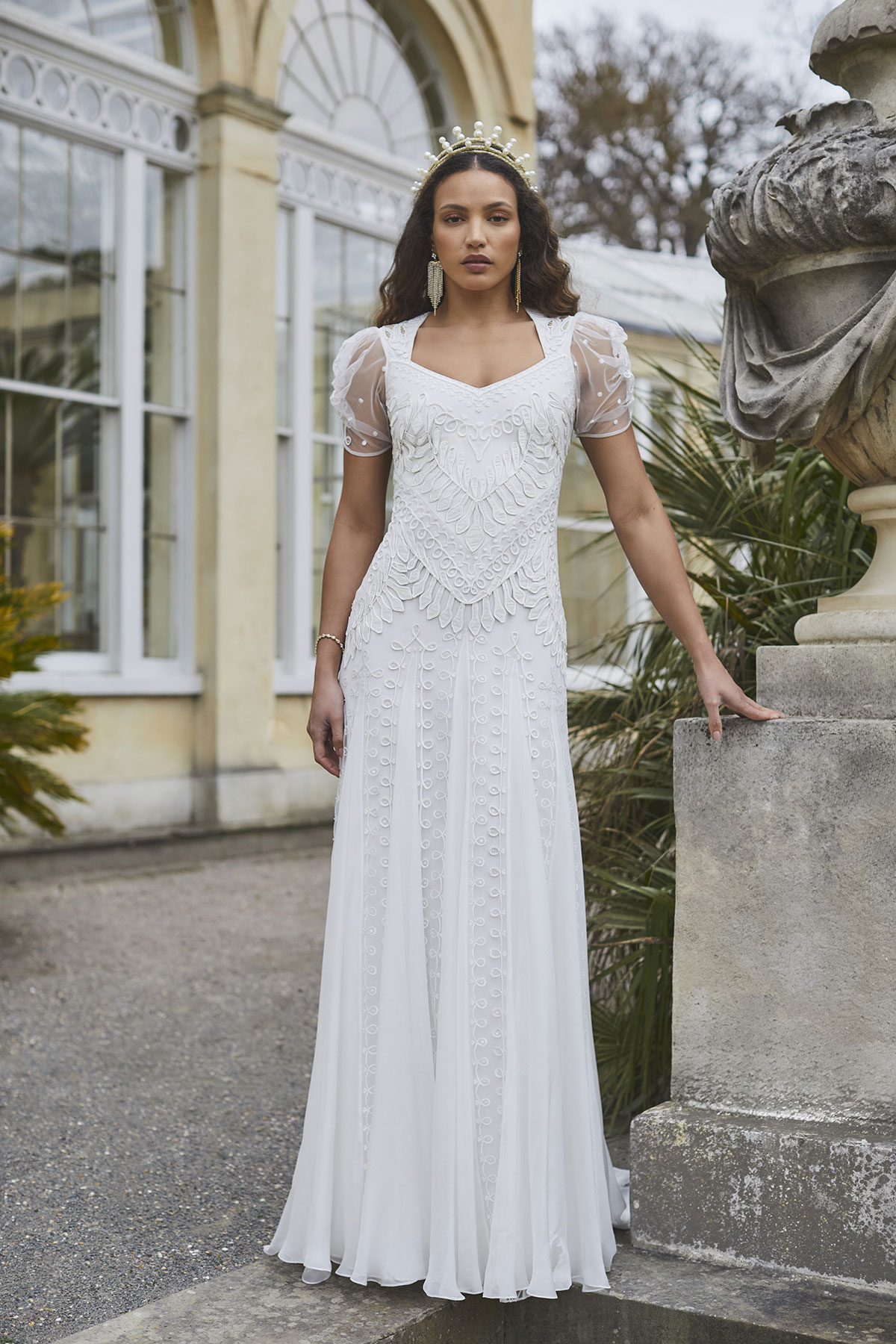 brunette model in short puff sleeve white dress, statement dangly earrings, and pearl tiara headband stands outside by stone structure