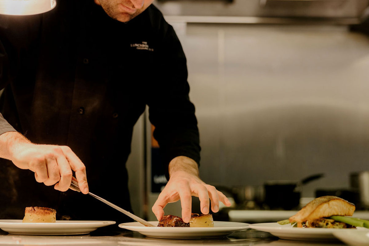 Chef plating a main course in a professional kitchen, arranging meat and vegetables on white dishes