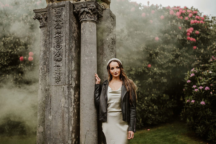 Bride wearing black leather jacket over her wedding dress outside in misty conditions