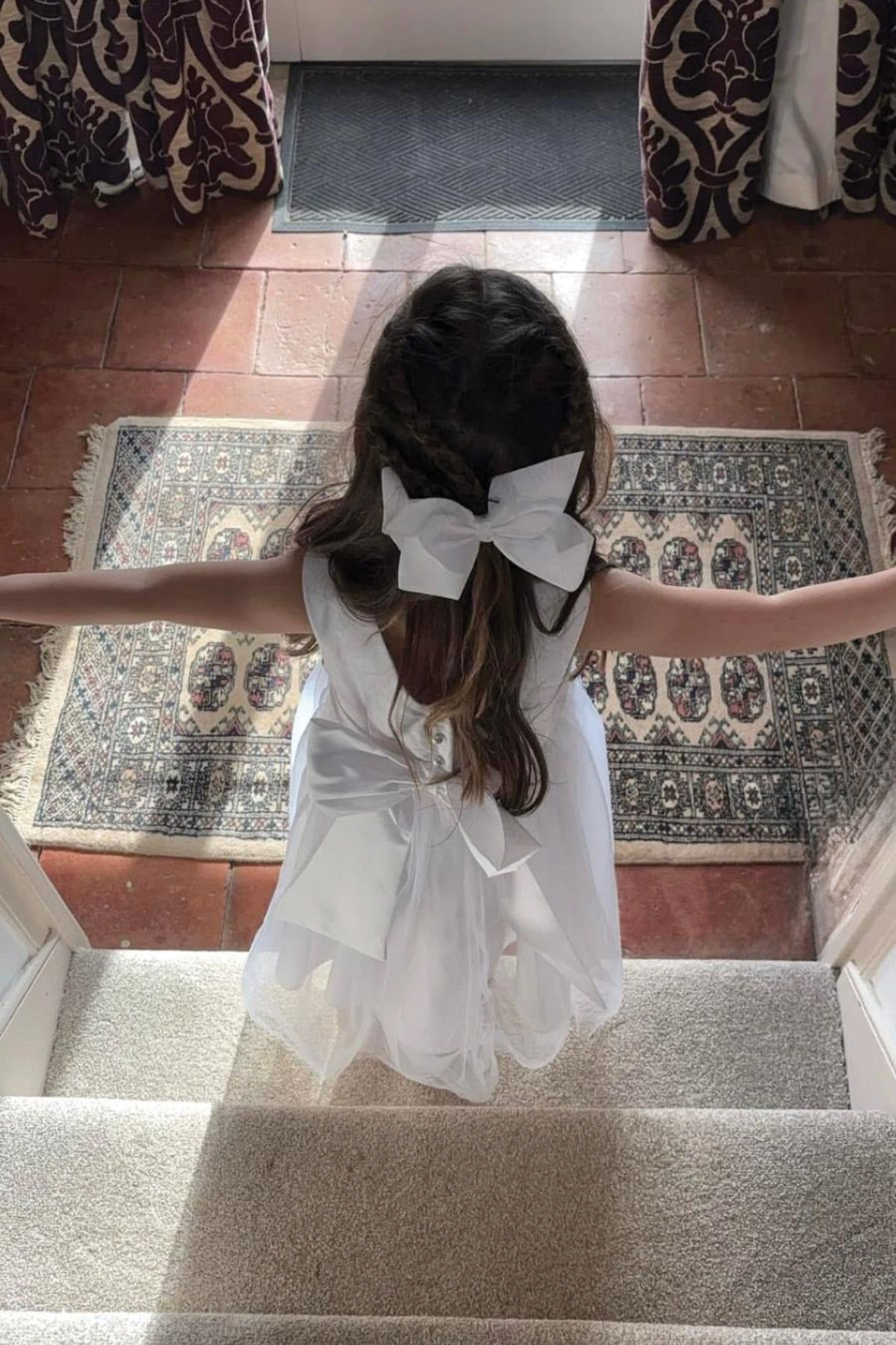 little girl in white dress with white bow on back and in her hair walks down carpet stairs