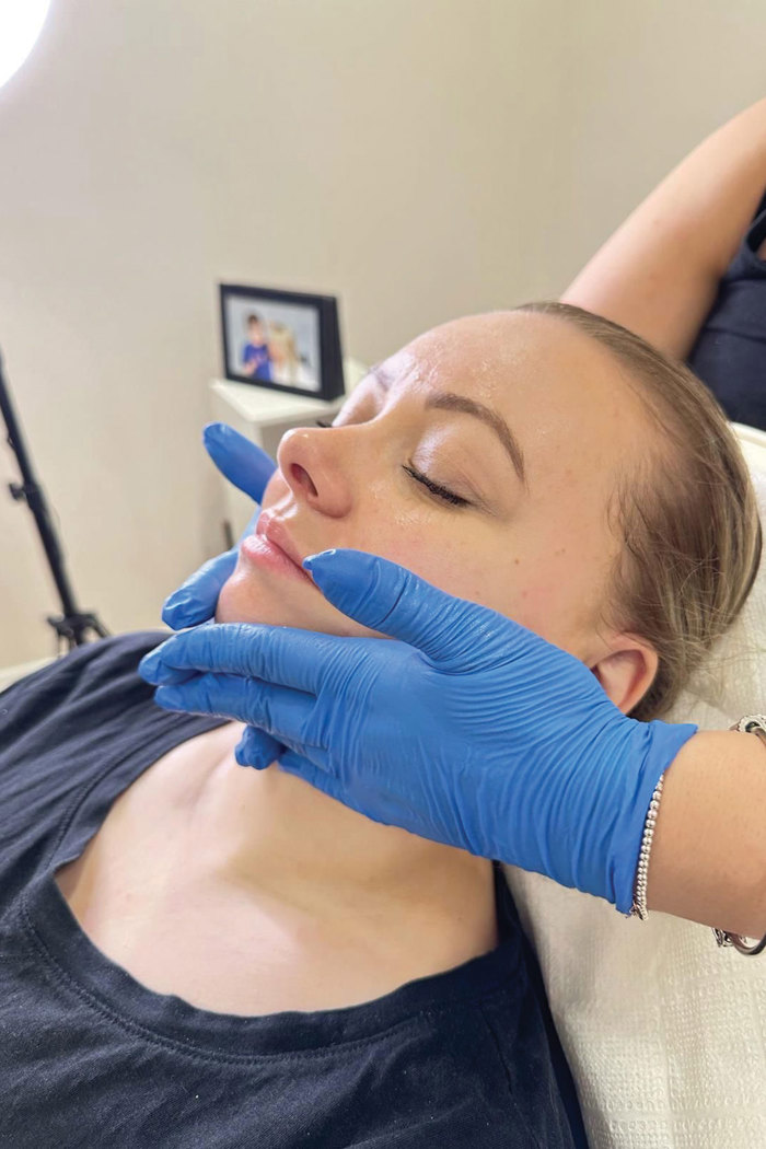 a blonde haired woman sat on a towel covered chair, having her face massaged by another person's blue medical glove covered hands