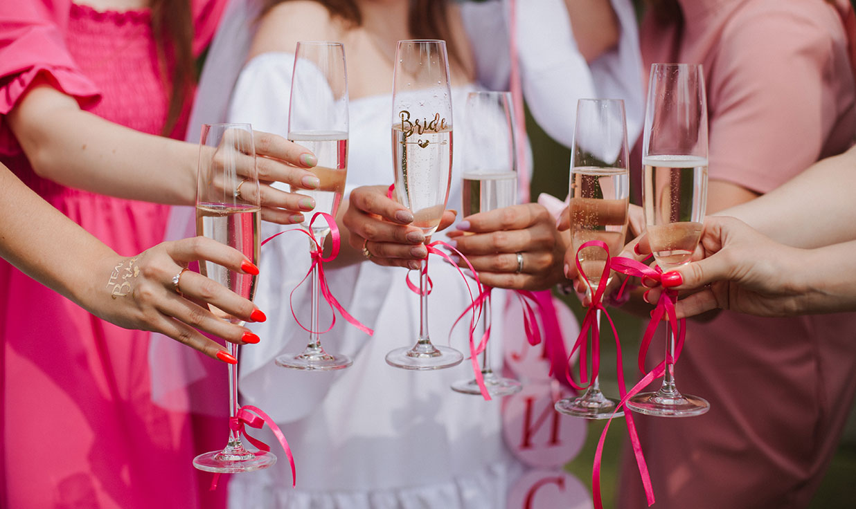bride in white and bridesmaids in pink all holding out glasses of champagne with pink ribbons tied around the stems and saying cheers