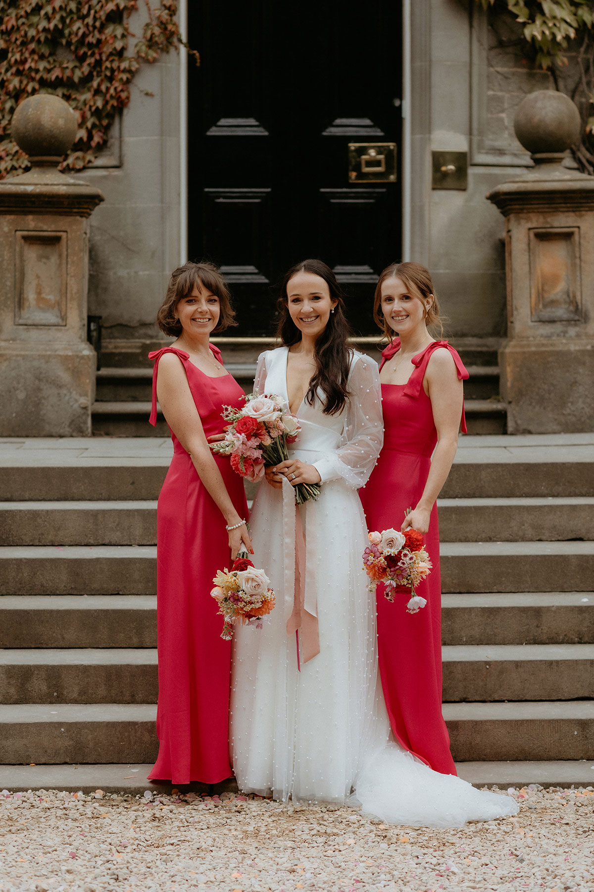 a bride posing with two bridesmaids in red dresses next to exterior stairs at Carlowrie Castle