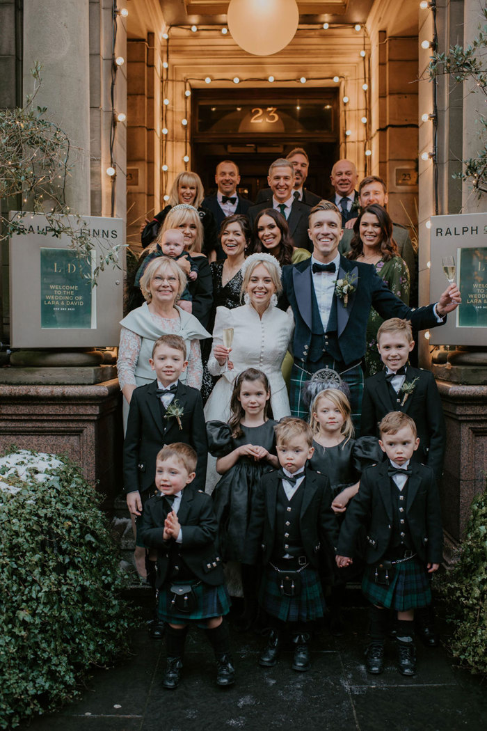 A group of people standing on an outdoor staircase in front of a doorway wearing dresses and kilts