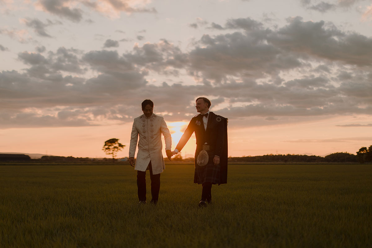 Newlywed grooms holding hands during golden hour portraits in open fields near Falkirk wedding venue
