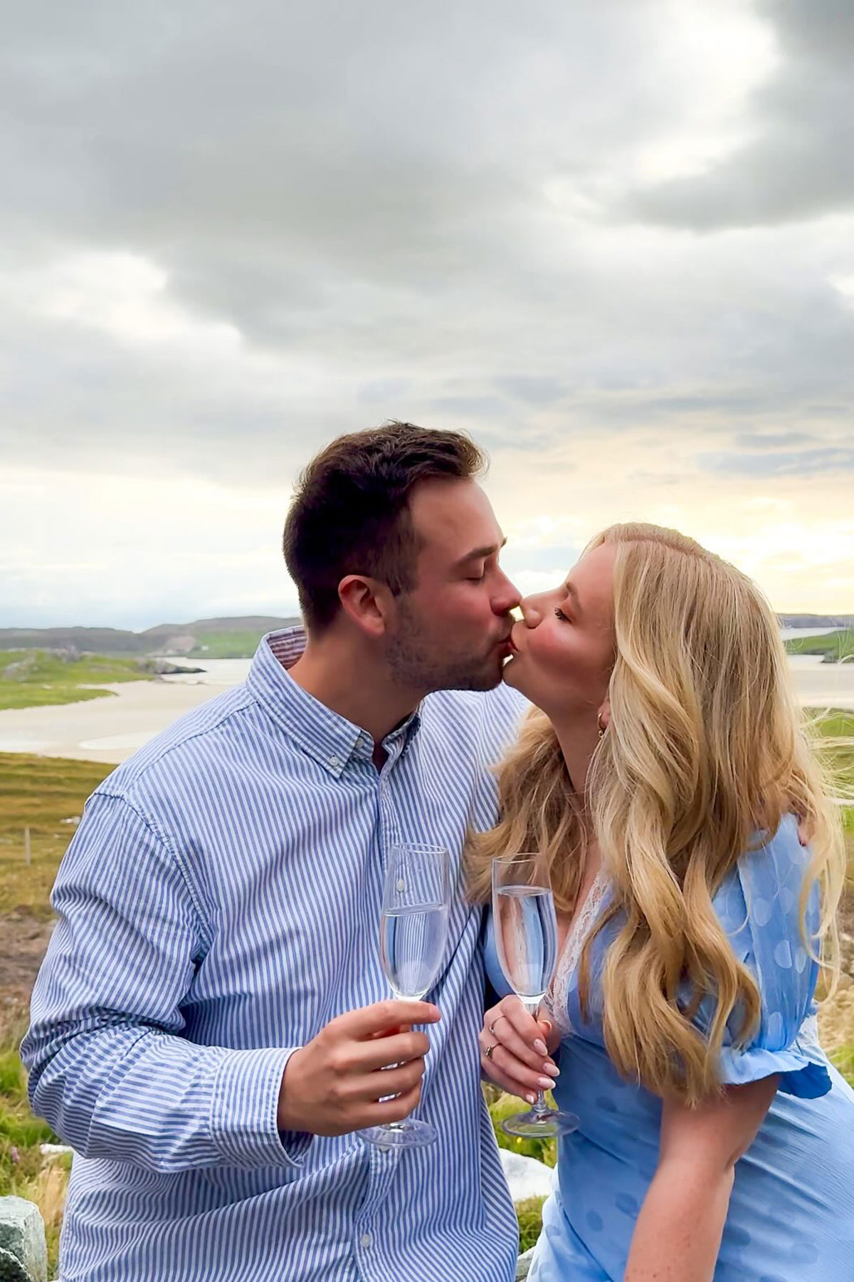 Engaged couple kissing while holding Champagne glasses against a dramatic Scottish coastal landscape at sunset