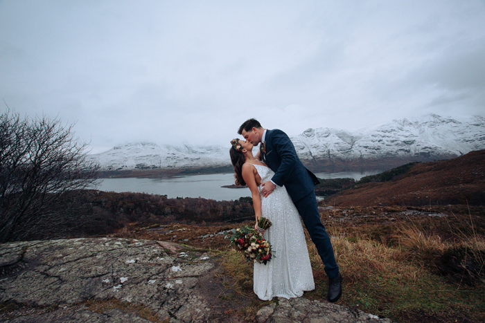Bride and groom kiss in front of loch with snow-capped mountains in the background