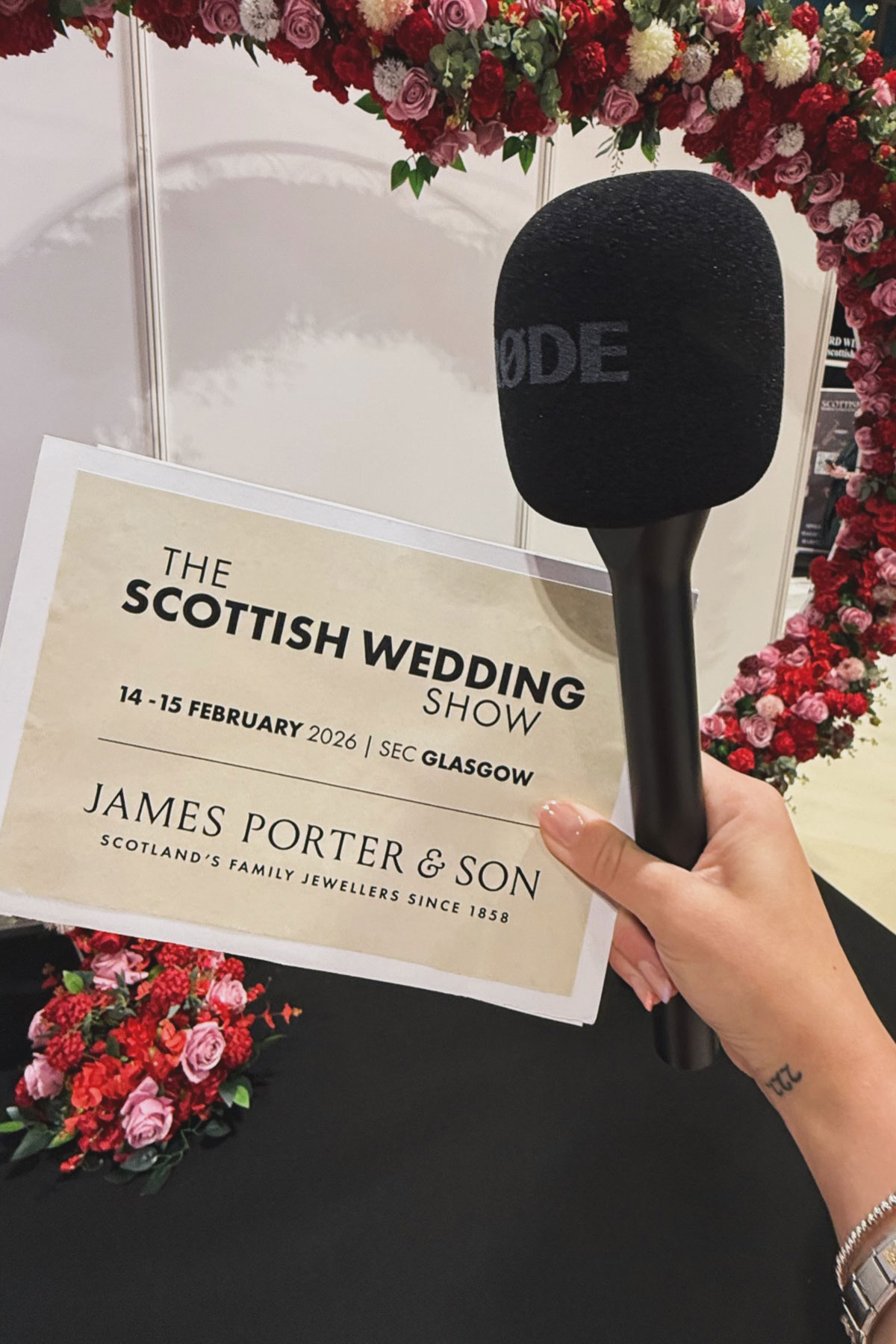 hand holding microphone and flyer for the scottish wedding show glasgow with floral display backdrop