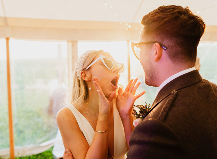 a bride and groom wearing heart shaped sunglasses in a light filled marquee with clear windows