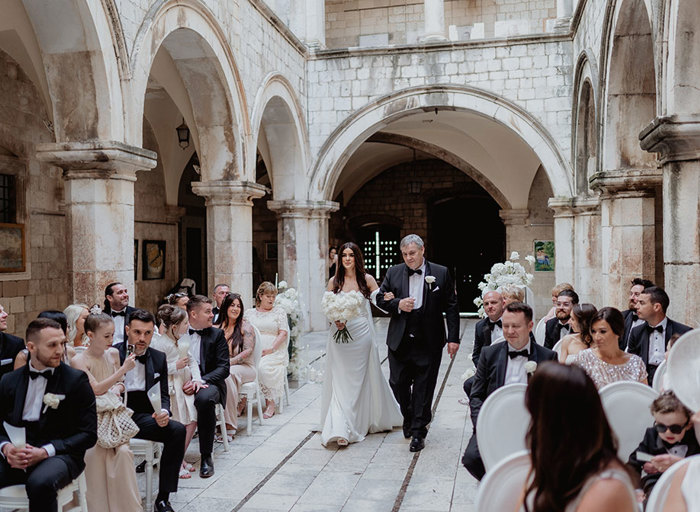 a bride walking down the aisle on the arm of a man wearing formal attire during a wedding ceremony at Sponsa Palace Dubrovnik