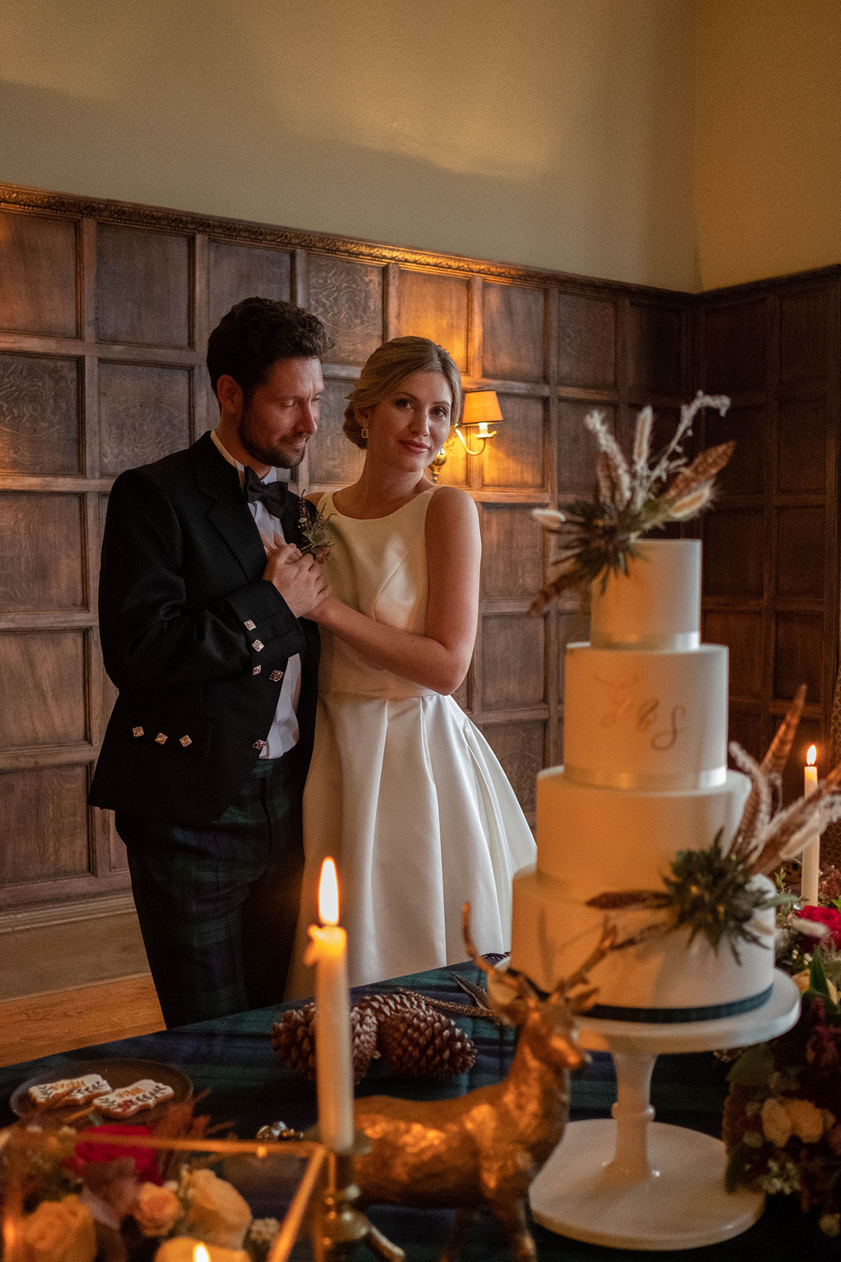 Bride and groom standing beside a three-tier wedding cake in a wood-panelled room, with candlelight, florals and a decorative stag ornament on the table