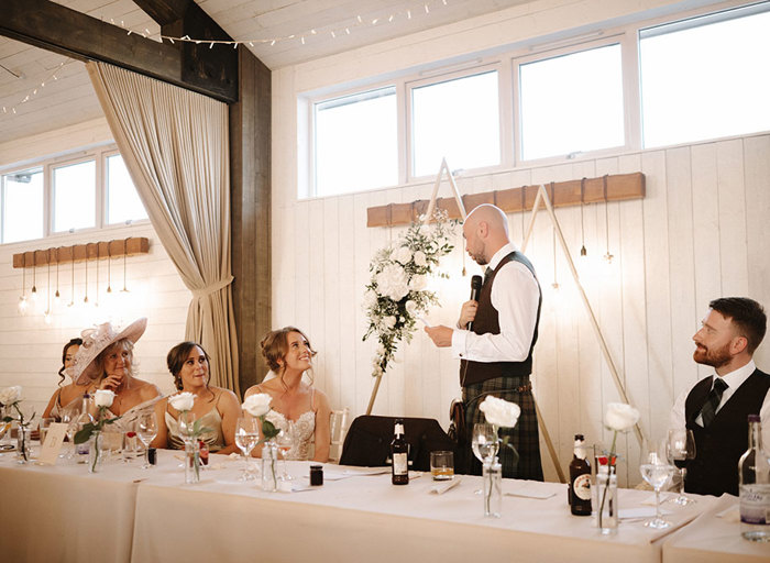 a groom stands holding a microphone while making a wedding speech looking towards a bride. Four other guests seated at the long top table look on 