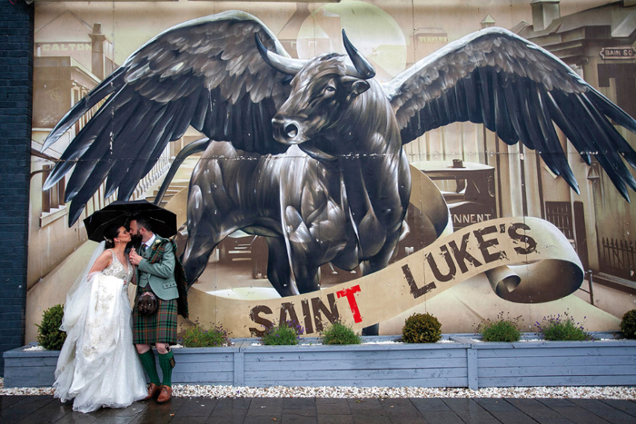 A bride and groom stand under an umbrella outside Saint Luke's