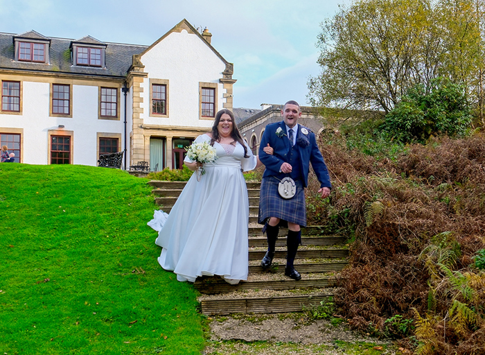 A bride in a flowy wedding dress and a groom in a blue kilt walk down stone steps in a garden with a building behind them]