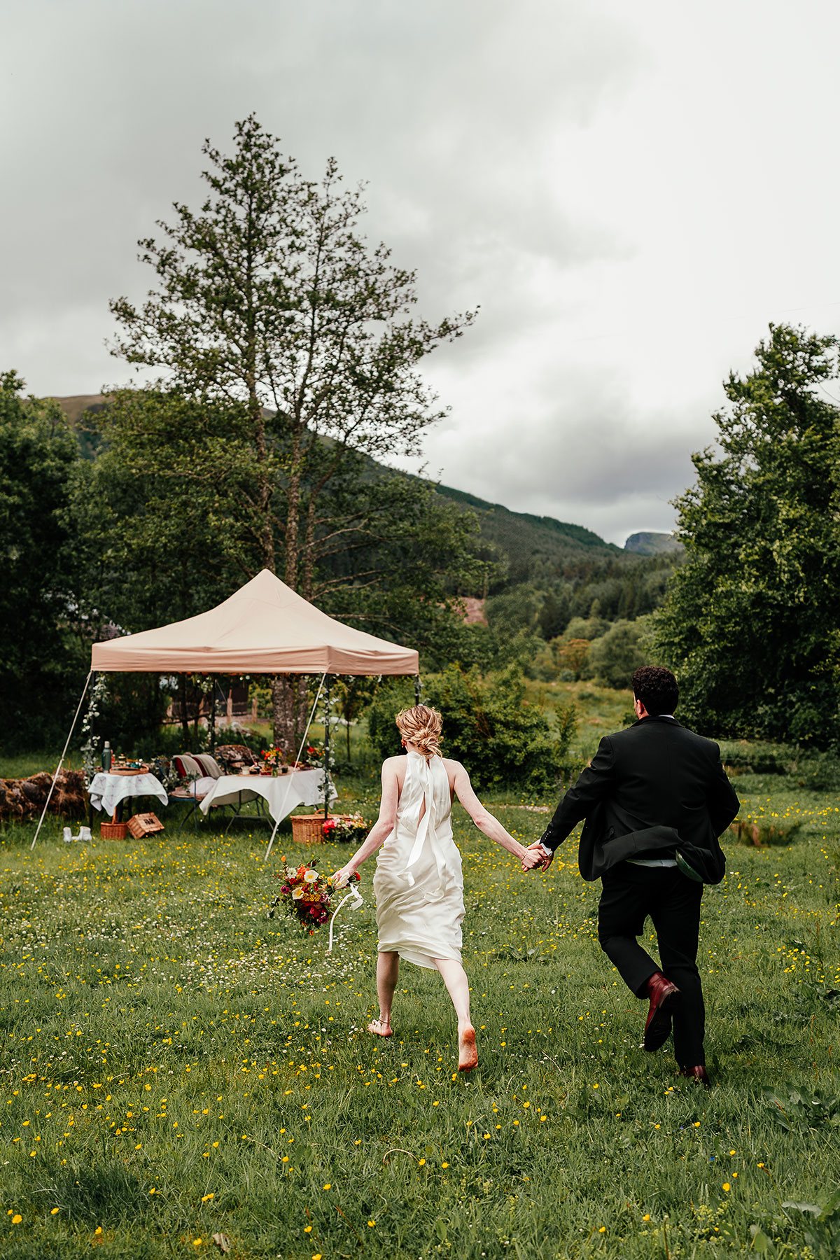 Newlyweds run hand in hand across a wildflower meadow towards an outdoor picnic setup in the Scottish countryside