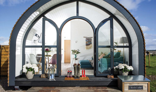 the front view of a pod with windows making up the entire front, including the door, has several pairs of wellies sat in front and a living and dining room visible within