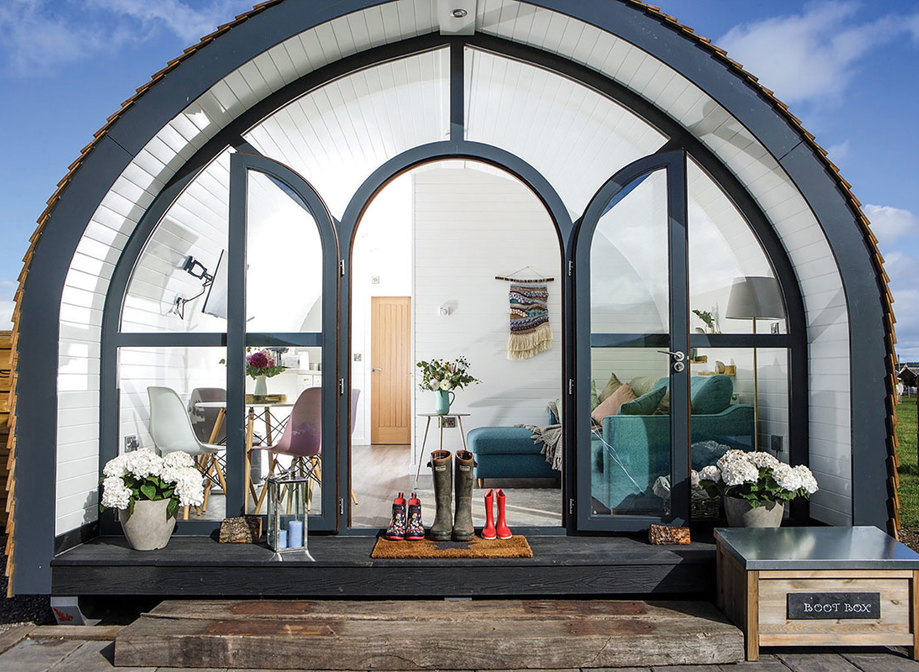 the front view of a pod with windows making up the entire front, including the door, has several pairs of wellies sat in front and a living and dining room visible within