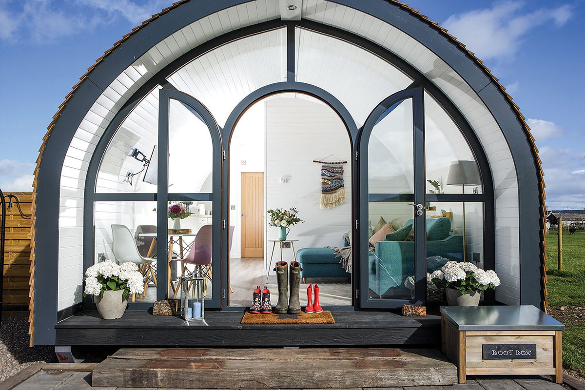 the front view of a pod with windows making up the entire front, including the door, has several pairs of wellies sat in front and a living and dining room visible within