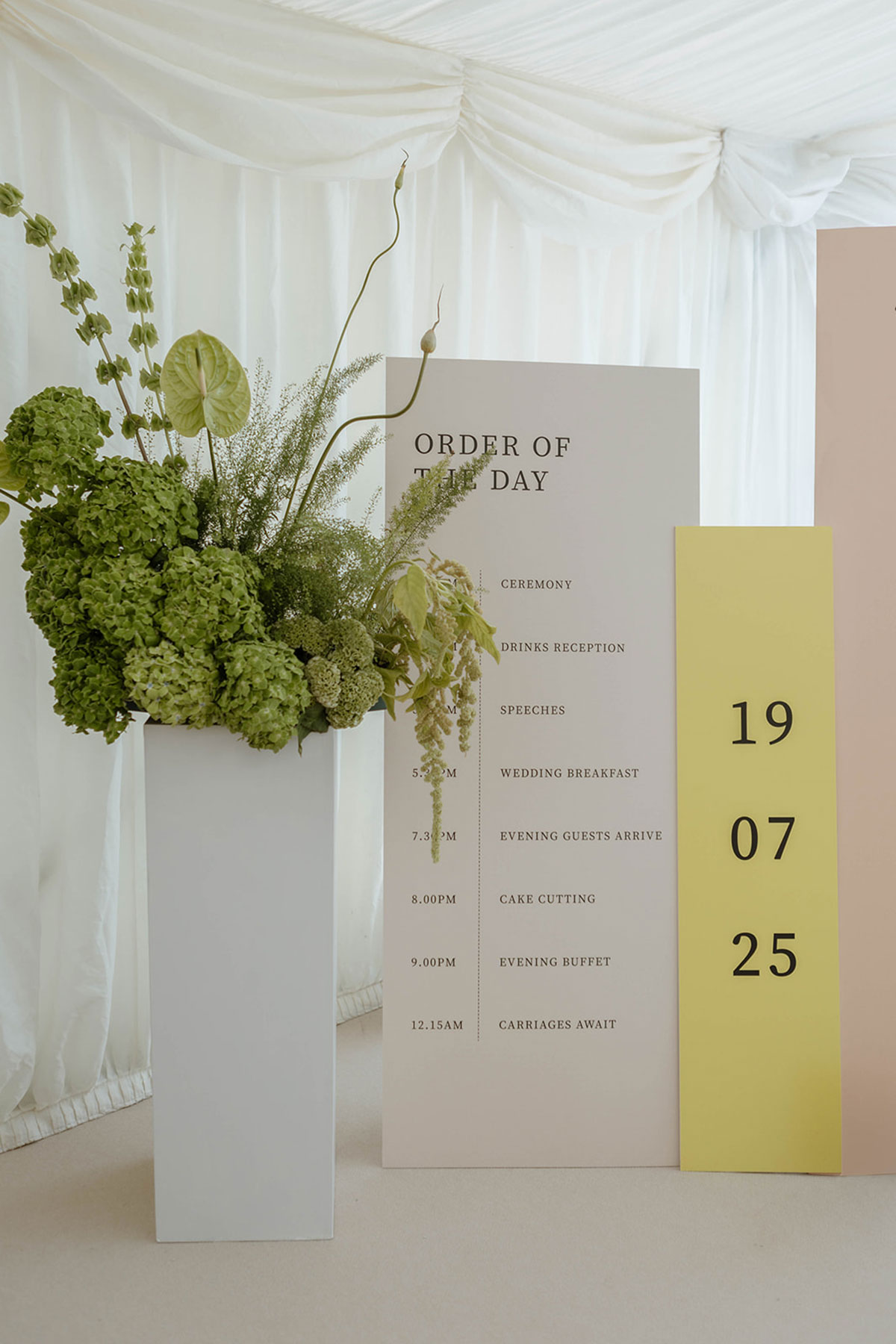 Wedding order of the day sign with green floral arrangement in a marquee reception space.