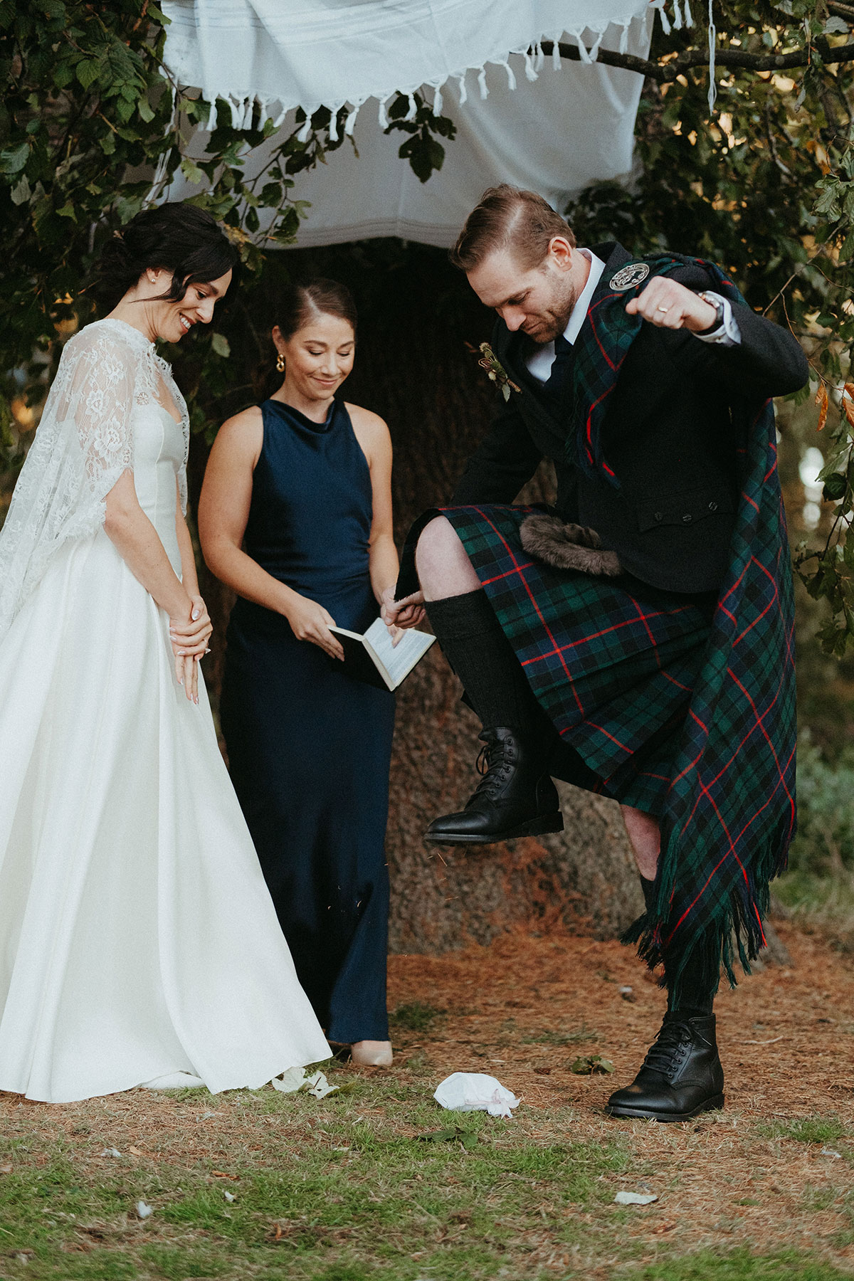 Groom takes part in the Jewish glass-smashing tradition beneath the chuppah as the bride and celebrant smile during the outdoor Cambo Country House ceremony.