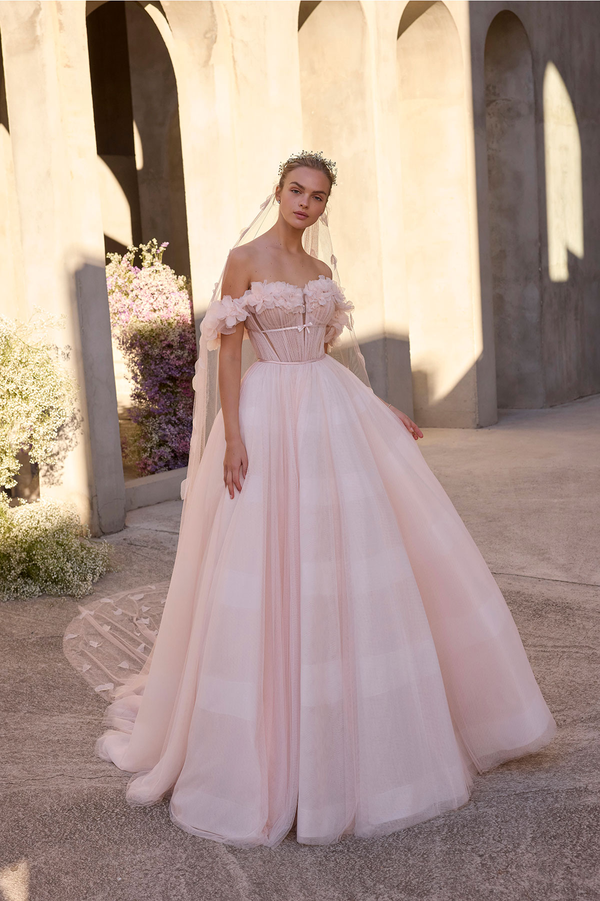 Bride wearing a blush pink off-the-shoulder ballgown wedding dress with ruffled neckline, corset bodice and full tulle skirt, photographed in warm light beneath stone arches
