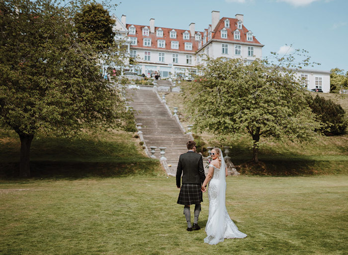 A groom in a kilt and a bride in a white dress stand on a grass lawn at the bottom of a tall set of stone stairs leading up to a large white building with an orange roof