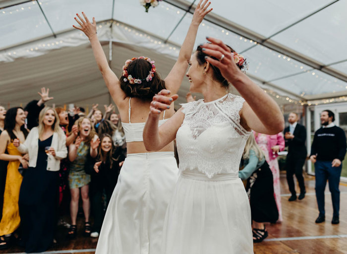 two brides throwing the bouquet in a marquee with surprised guests looking animated, some with hands outstretched trying to catch it