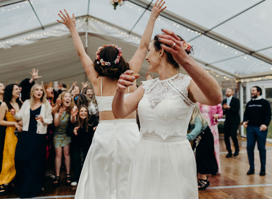two brides throwing the bouquet in a marquee with surprised guests looking animated, some with hands outstretched trying to catch it