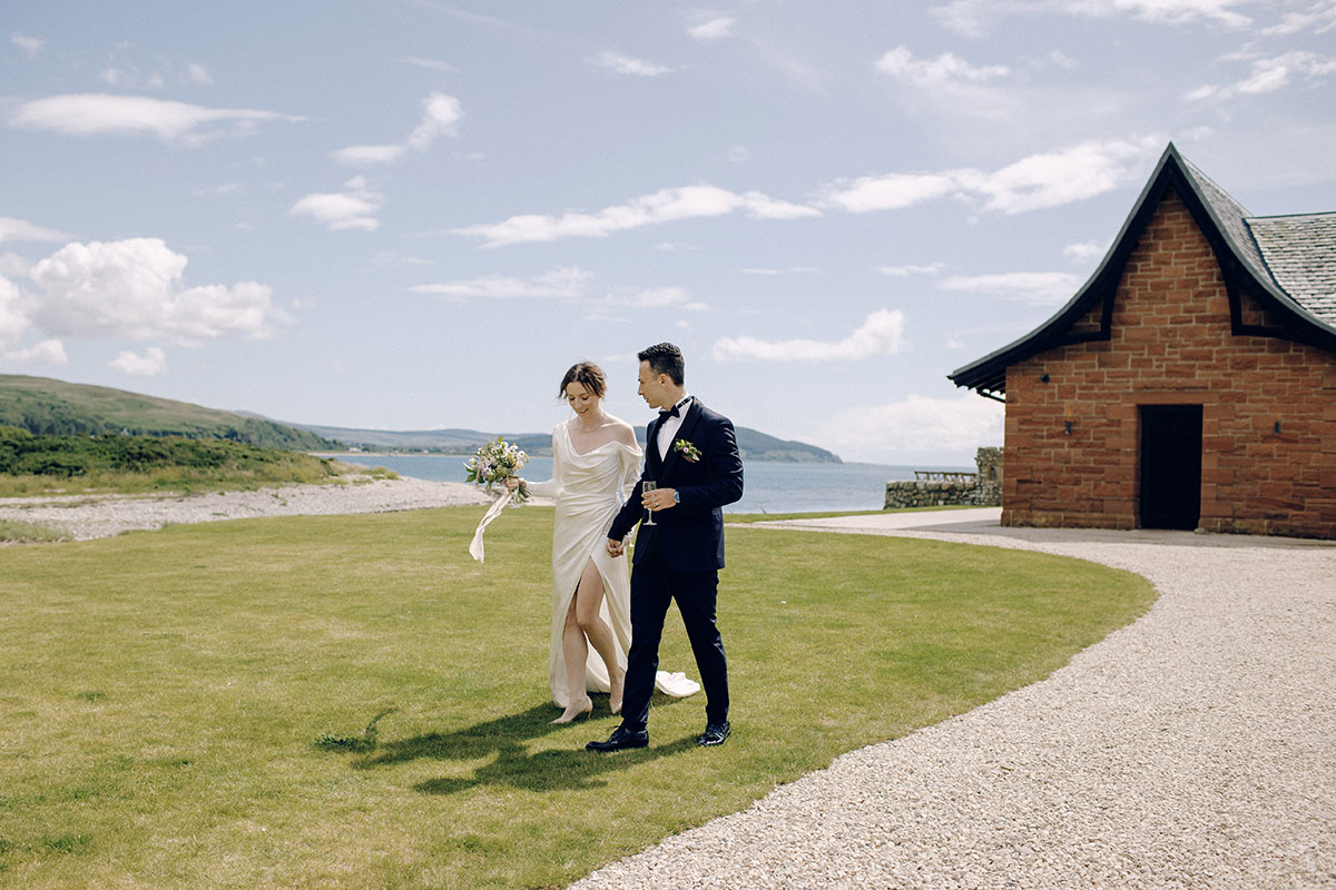 bride in silky cream wedding dress and and groom in black tuxedo hold hands and walk across a green patch of grass with a brick boathouse and blue skies and water visible in the close background
