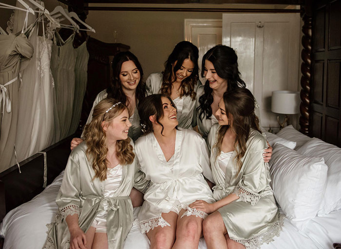 Six women sitting on a bed, one in a white silk robe and the others in sage green silk robes