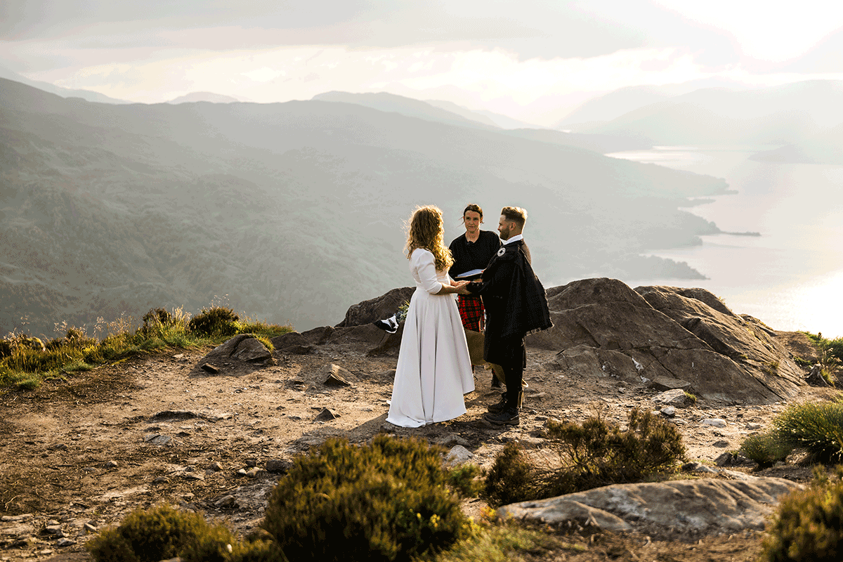 Couple stand with their celebrant during a mountaintop humanist wedding ceremony overlooking a Scottish loch at sunset