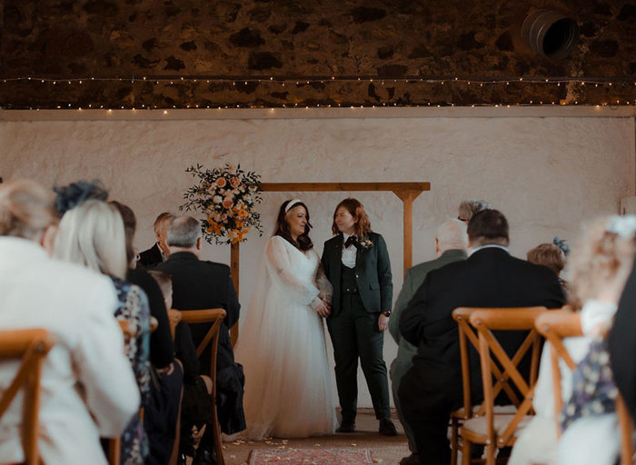 two brides holding hands at the end of a wedding ceremony at Cow Shed Crail. They stand below a wooden arch with a flower posy on the top left corner and rows of wedding guests sitting on wooden cross back chairs look on