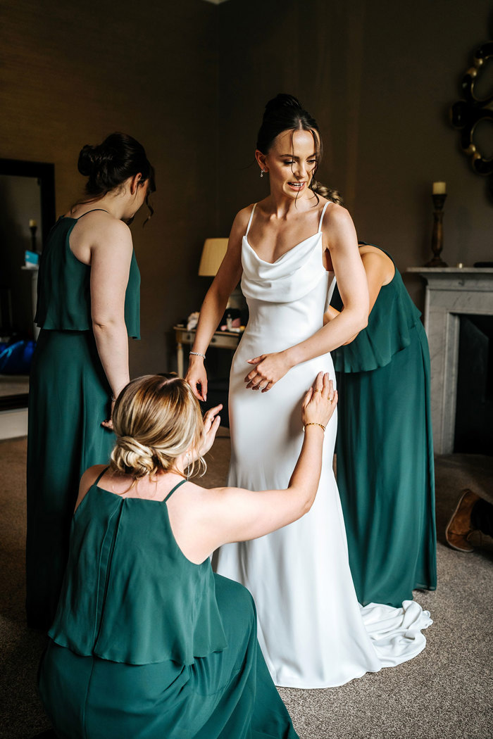 three bridesmaids wearing floor length green dresses help bride into wedding dress