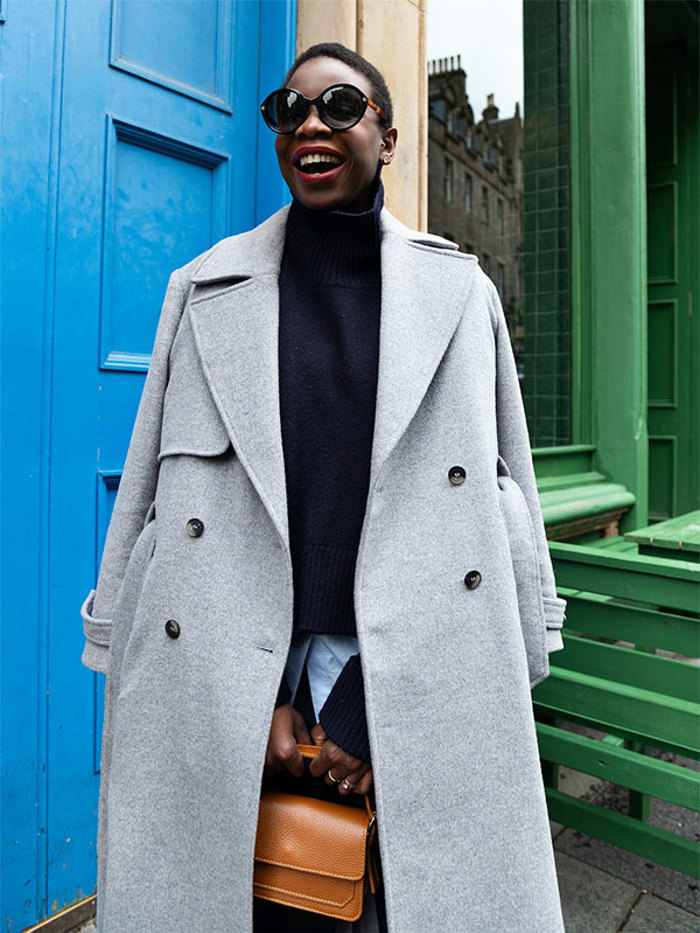 a woman smiles outside a green and blue building wearing red lipstick, a long grey coat and a black rollneck jumper