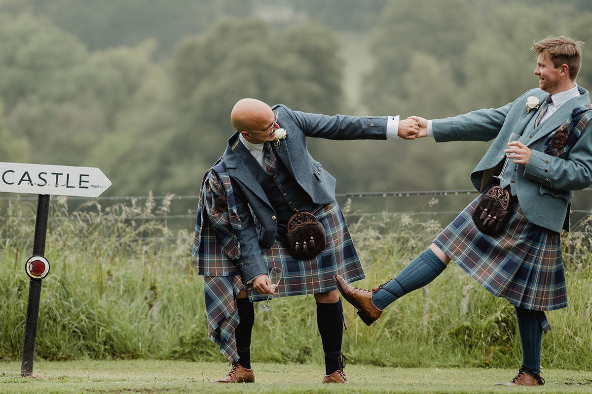 The grooms share a playful moment outdoors, holding hands and laughing near a sign pointing towards a castle