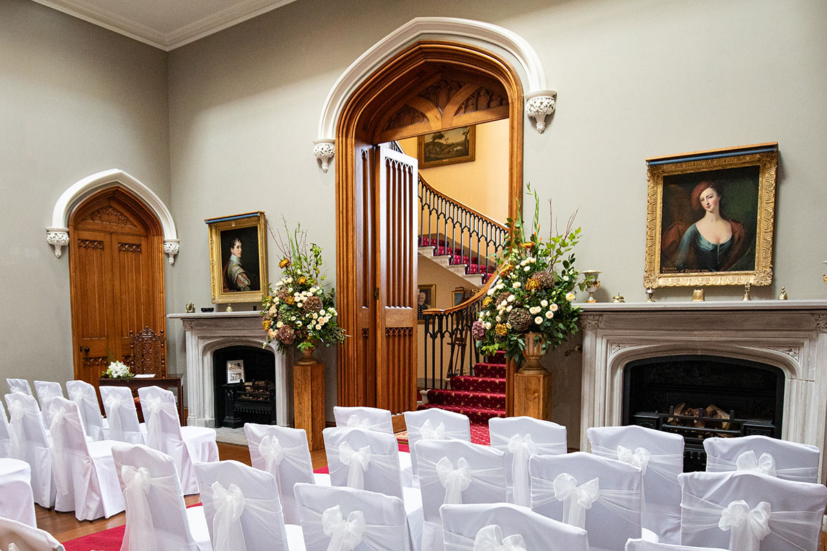 Wedding ceremony setup inside Blairquhan Castle’s saloon, with white chair covers, floral displays and portraits framing the grand staircase.