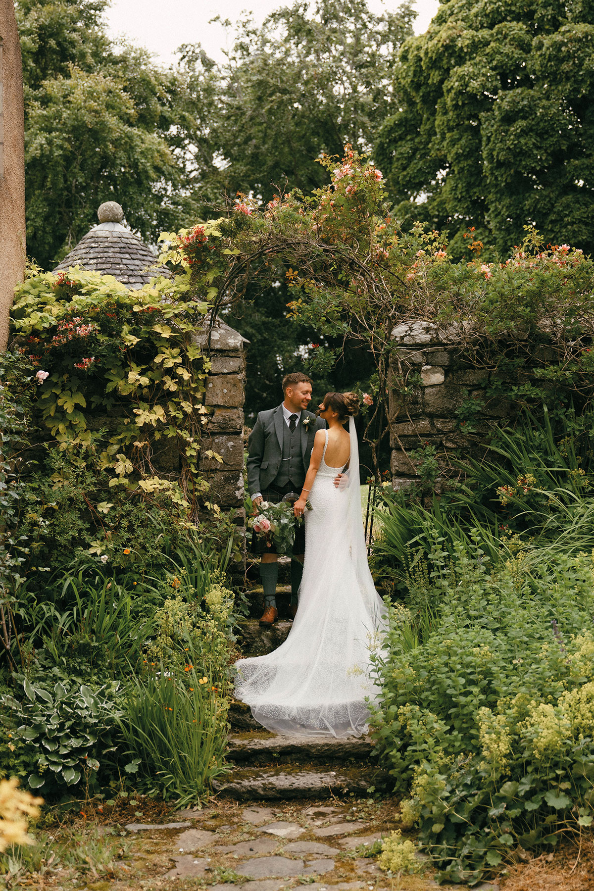Bride and groom standing together beneath a flower-covered stone archway surrounded by lush gardens at Aswanley wedding venue in Aberdeenshire