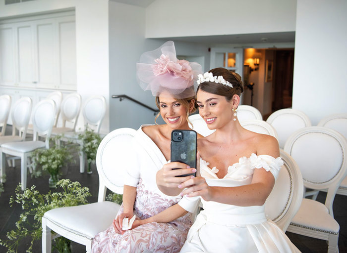 A woman in a white and pink dress and a pink fascinator sits next to a bride who is holding an iPhone and taking a selfie of them both