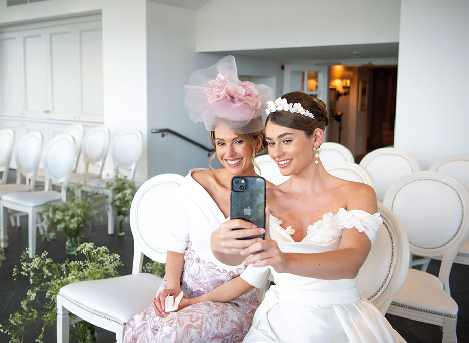 A woman in a white and pink dress and a pink fascinator sits next to a bride who is holding an iPhone and taking a selfie of them both