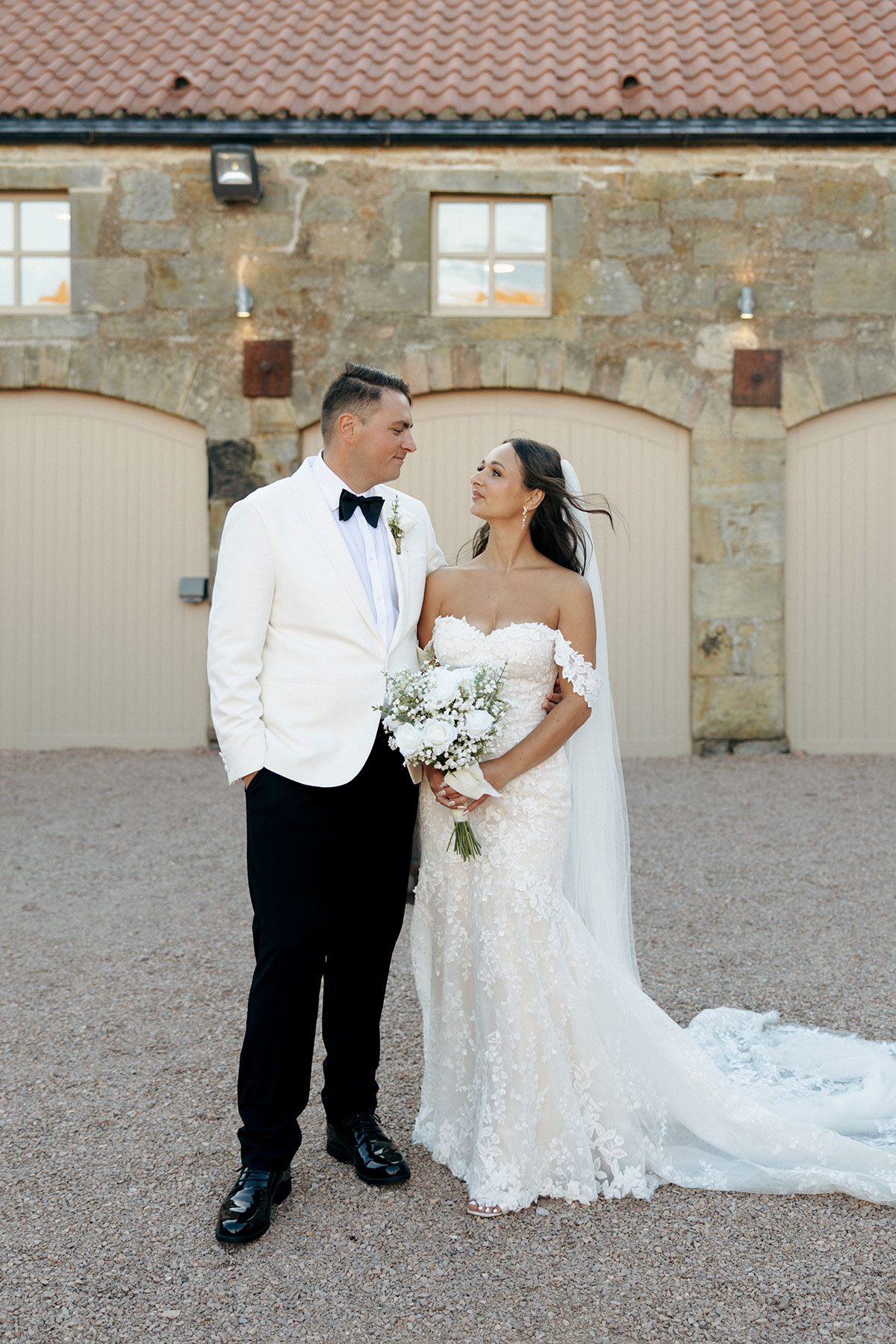 Newlyweds pose together in the courtyard at Falside Mill, with soft evening light and the stone barn doors behind them.