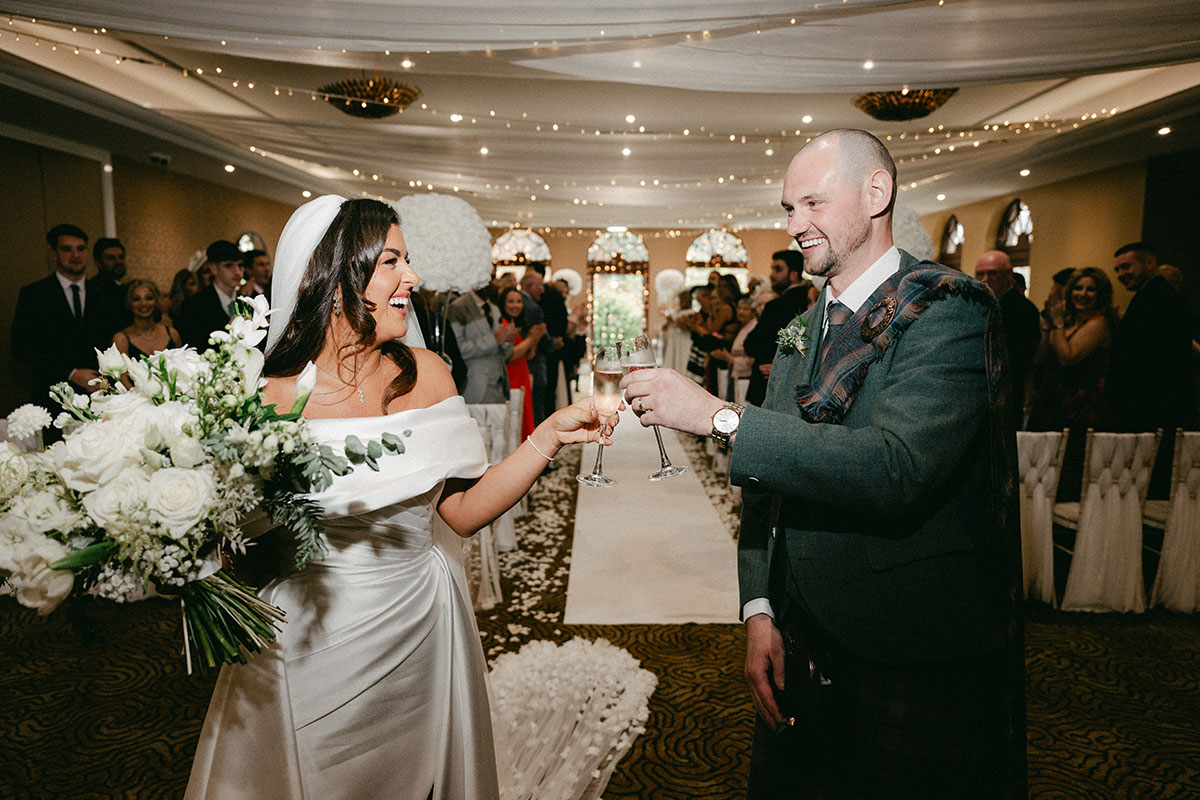 bride in off the shoulder wedding dress holds a large white bouquet and cheers a champagne glass at top of aisle with groom in green, blue and purple tartan kilt outfit with green jacket